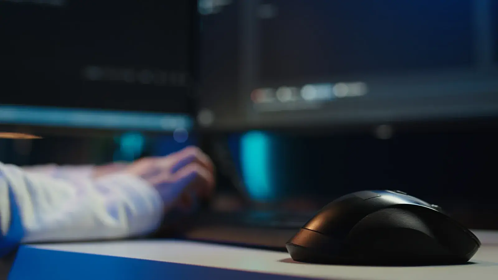 Hand an der Tastatur vor unscharfen Bildschirmen mit Code, im Vordergrund eine Computermaus, in blauem Licht