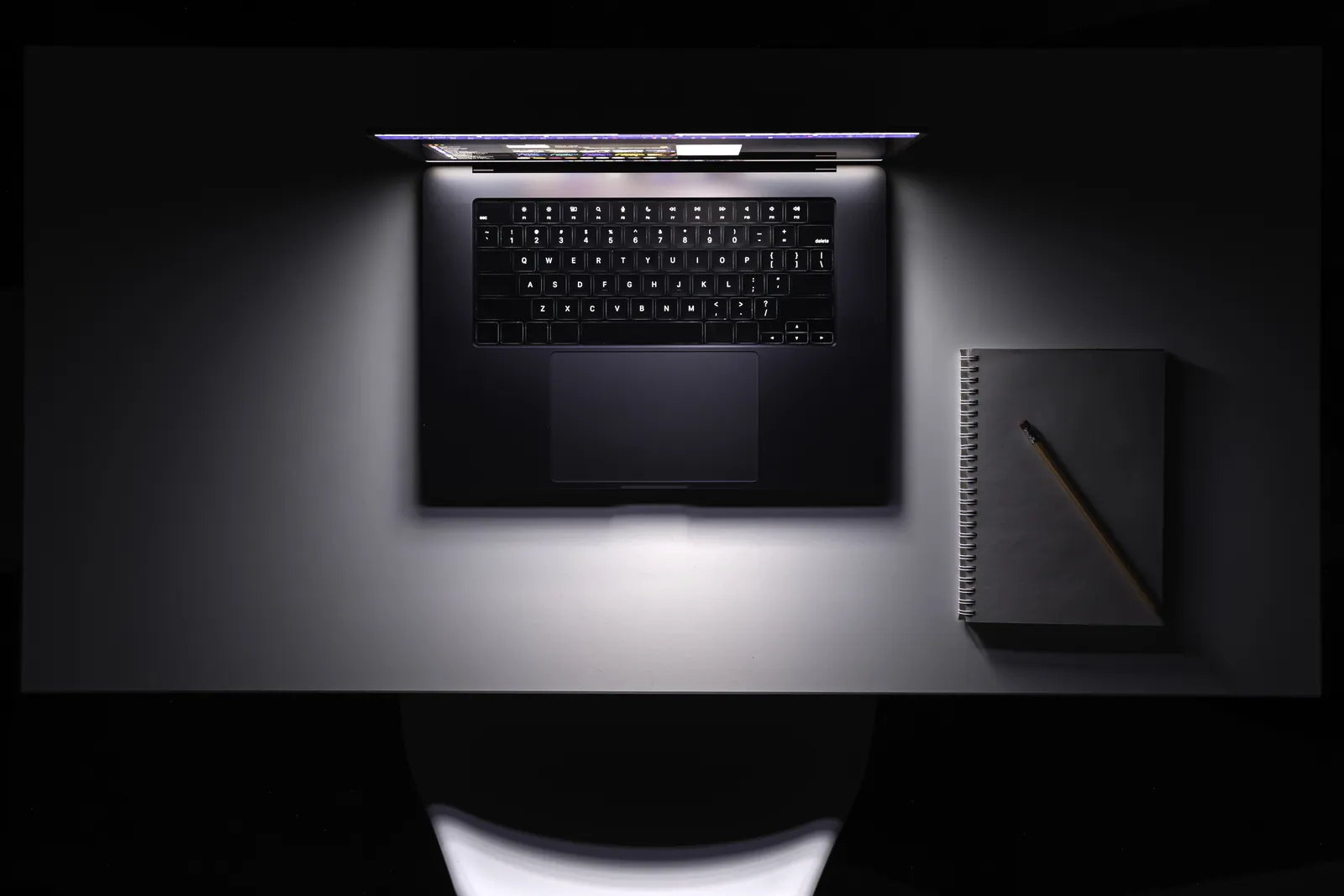 Aufgeklappter Laptop mit beleuchteter Tastatur neben einem Notizbuch mit Stift auf einem dunklen Tisch
